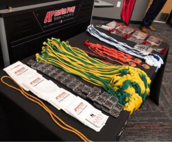 Table with award medals on green and yellow cords, red/white/blue lanyards, and white drawstring bags at a university event with branding in the background.