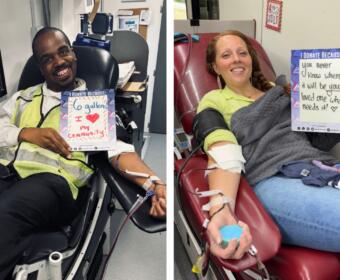 Two blood donors in a clinic, smiling and holding signs about why they donate.