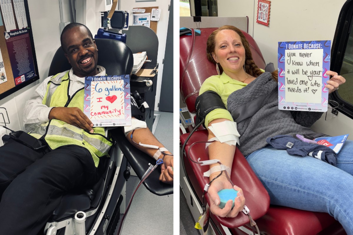 Two blood donors in a clinic, smiling and holding signs about why they donate.