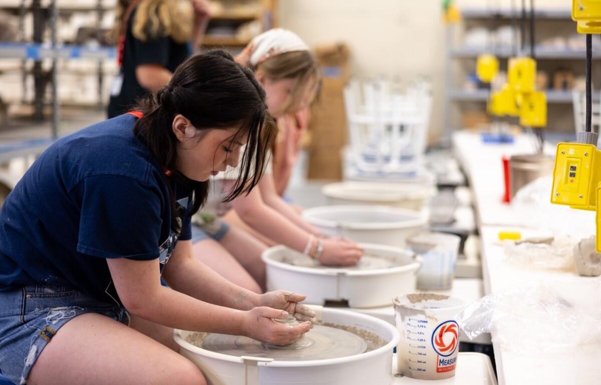 Teen girl shaping clay on a pottery wheel in a classroom, with other students working at wheels in the background.