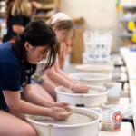 Teen girl shaping clay on a pottery wheel in a classroom, with other students working at wheels in the background.