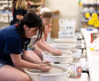 Teen girl shaping clay on a pottery wheel in a classroom, with other students working at wheels in the background.