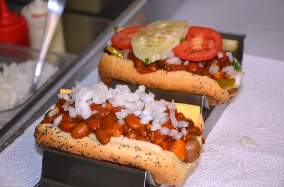 Three chili dogs in poppy seed buns topped with beans, diced onions, jalapeños, tomato slices, a pickle, and a single potato chip on top, resting in a metal bun steamer.