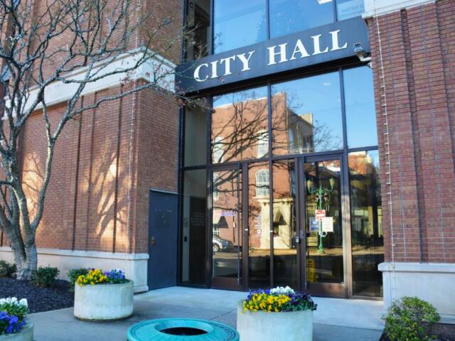 City Hall entrance with large glass doors, brick exterior, and colorful flower planters along the walkway.