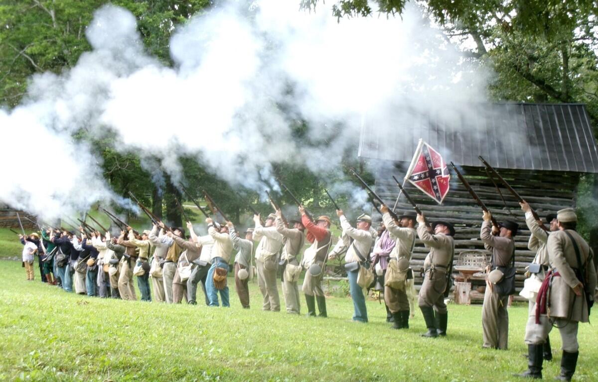 Line of Civil War reenactors firing muskets, smoke rising from barrels in a grassy field with trees behind.