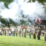 Line of Civil War reenactors firing muskets, smoke rising from barrels in a grassy field with trees behind.