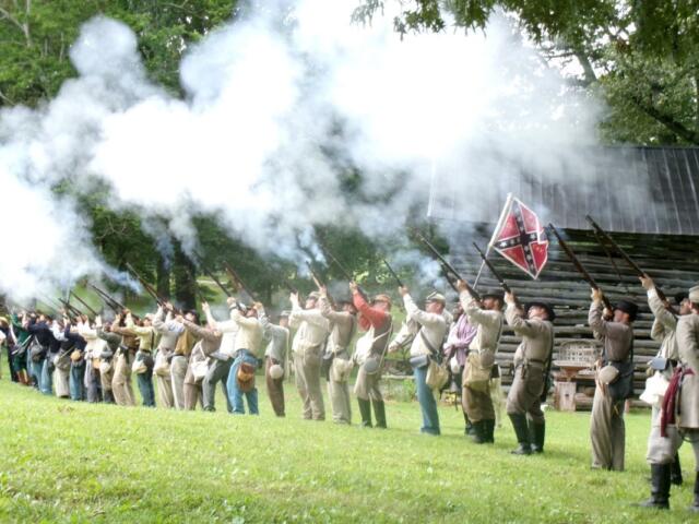 Line of Civil War reenactors firing muskets, smoke rising from barrels in a grassy field with trees behind.
