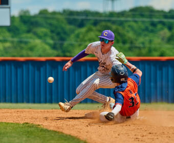 Baseball scene: runner in orange and blue slides into a base while the fielder in pinstripe attempts a tag; ball is bouncing nearby on the dirt.