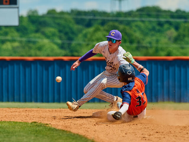 Baseball scene: runner in orange and blue slides into a base while the fielder in pinstripe attempts a tag; ball is bouncing nearby on the dirt.