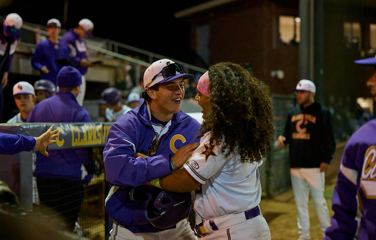 Two baseball players in purple and white uniforms hug and laugh in the dugout at night.