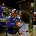 Two baseball players in purple and white uniforms hug and laugh in the dugout at night.