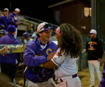 Two baseball players in purple and white uniforms hug and laugh in the dugout at night.