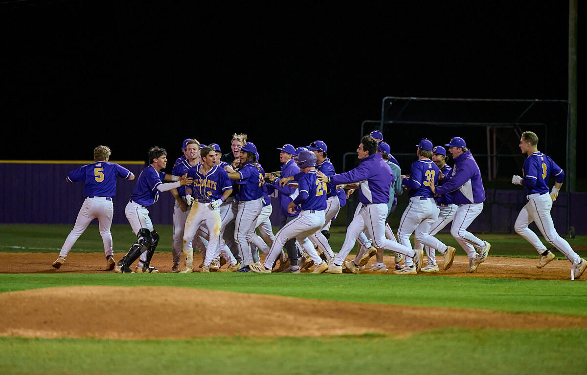 Baseball team in blue uniforms celebrates together on the infield at night, embracing and cheering near home plate.