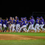 Baseball team in blue uniforms celebrates together on the infield at night, embracing and cheering near home plate.