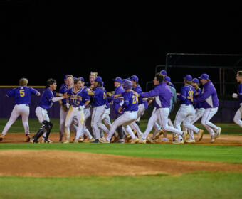 Baseball team in blue uniforms celebrates together on the infield at night, embracing and cheering near home plate.