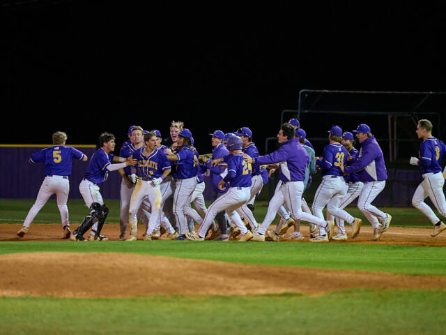 Baseball team in blue uniforms celebrates together on the infield at night, embracing and cheering near home plate.