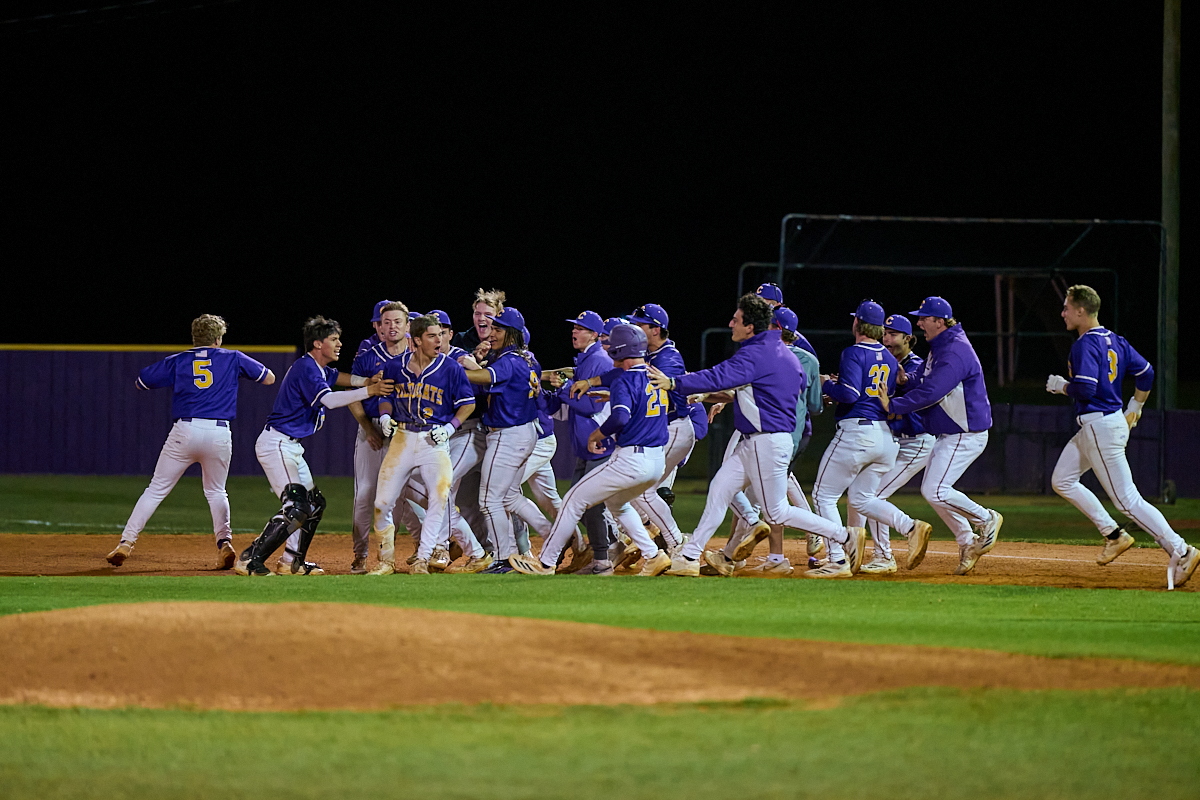 Baseball team in blue uniforms celebrates together on the infield at night, embracing and cheering near home plate.