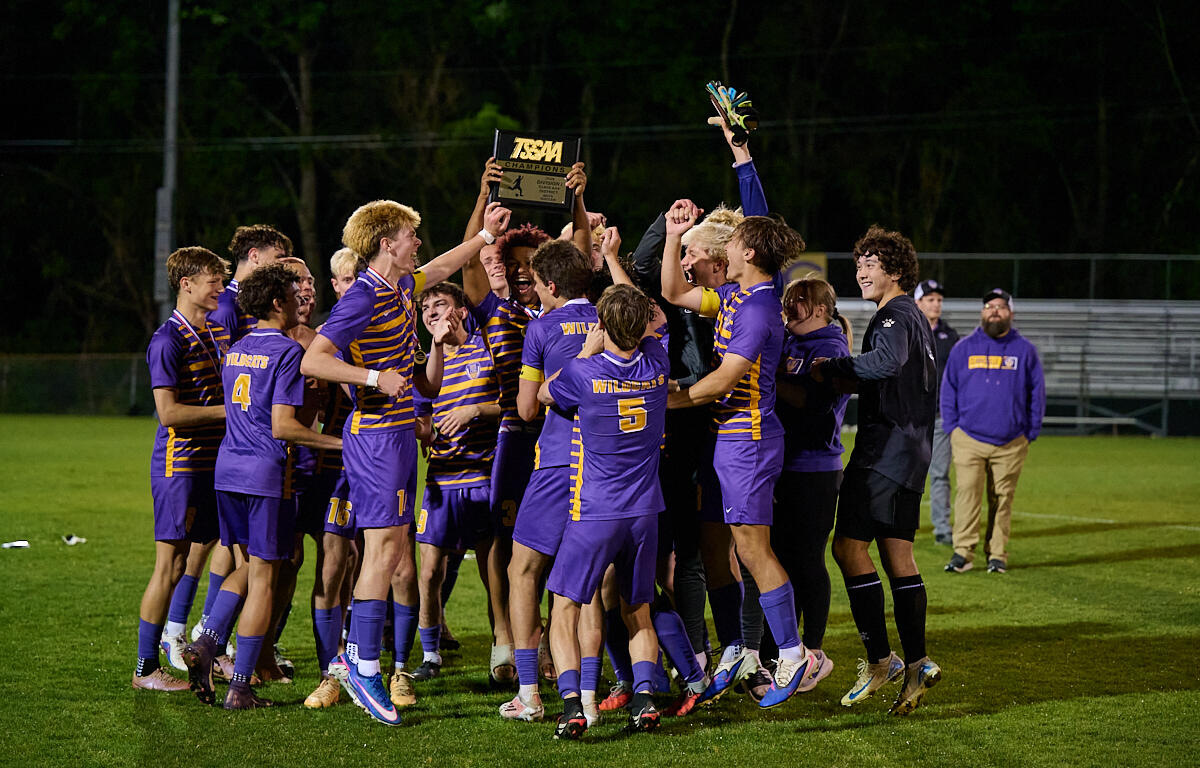 Purple and gold soccer team celebrates on a grassy field, lifting a championship trophy together.