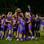 Purple and gold soccer team celebrates on a grassy field, lifting a championship trophy together.