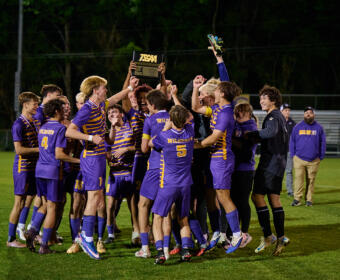 Purple and gold soccer team celebrates on a grassy field, lifting a championship trophy together.
