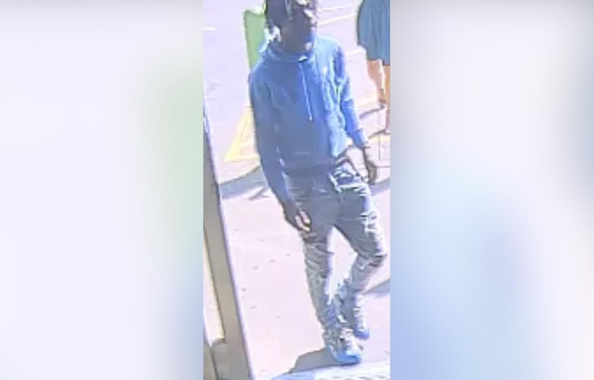 Person wearing blue hoodie, dark jeans, cap, and face mask standing outside a storefront. (CCTV-style image)