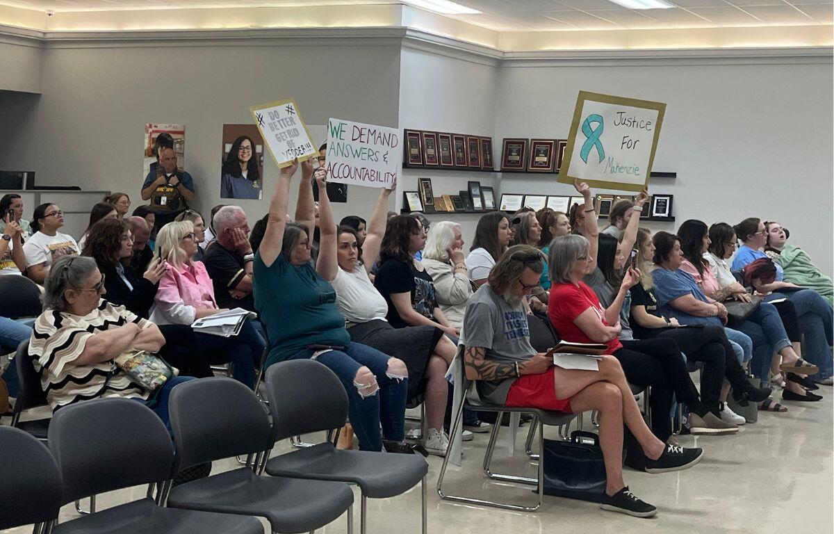People seated in a community meeting, several raising signs demanding accountability and justice.