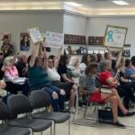 People seated in a community meeting, several raising signs demanding accountability and justice.