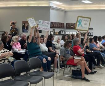People seated in a community meeting, several raising signs demanding accountability and justice.