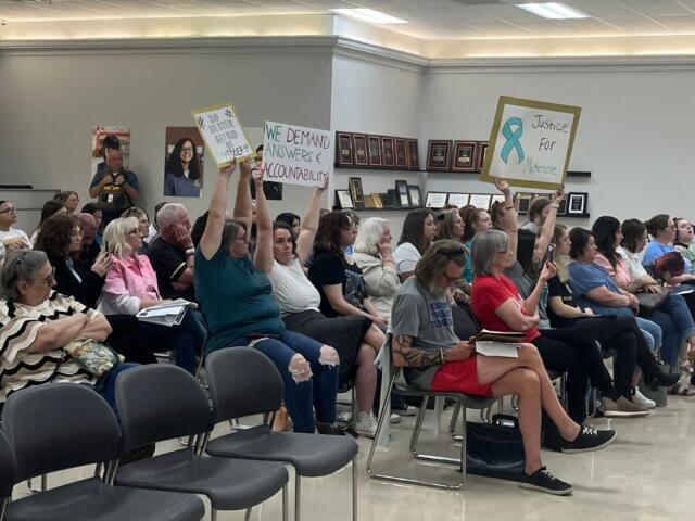 People seated in a community meeting, several raising signs demanding accountability and justice.