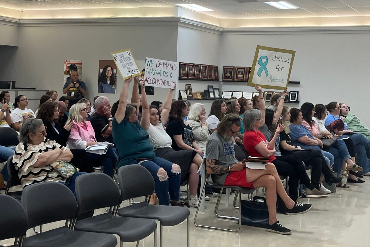 People seated in a community meeting, several raising signs demanding accountability and justice.
