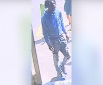 Person wearing blue hoodie, dark jeans, cap, and face mask standing outside a storefront. (CCTV-style image)