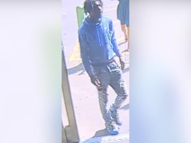 Person wearing blue hoodie, dark jeans, cap, and face mask standing outside a storefront. (CCTV-style image)