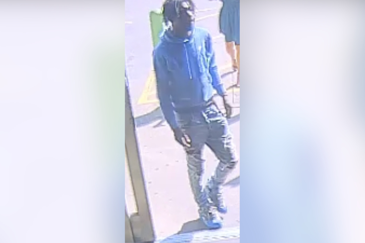 Person wearing blue hoodie, dark jeans, cap, and face mask standing outside a storefront. (CCTV-style image)