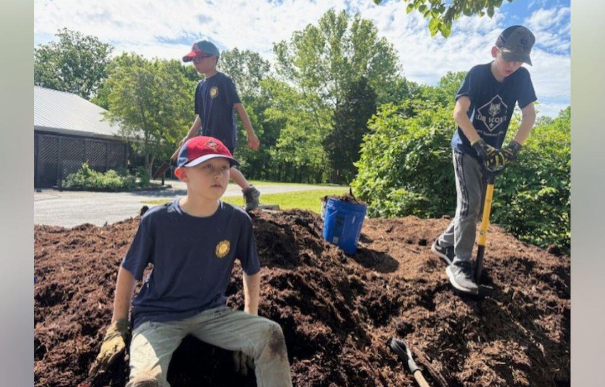 Three boys digging a large dirt mound outdoors on a sunny day, one seated while two stand with shovels nearby.