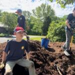 Three boys digging a large dirt mound outdoors on a sunny day, one seated while two stand with shovels nearby.