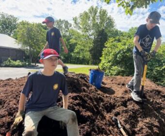 Three boys digging a large dirt mound outdoors on a sunny day, one seated while two stand with shovels nearby.