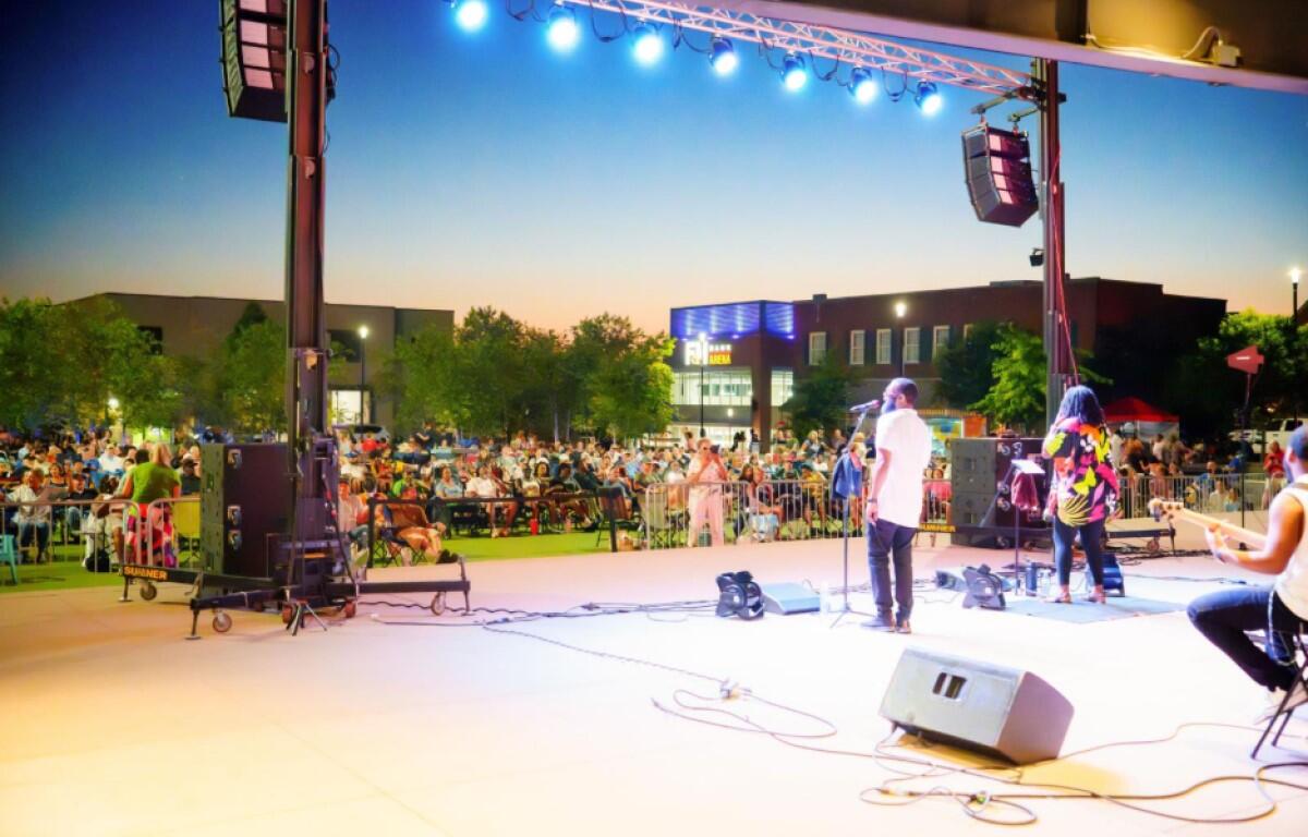 Outdoor concert at dusk with a band performing on stage and a large crowd seated beyond the barriers.