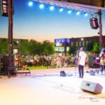 Outdoor concert at dusk with a band performing on stage and a large crowd seated beyond the barriers.