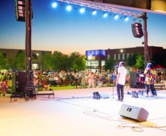 Outdoor concert at dusk with a band performing on stage and a large crowd seated beyond the barriers.