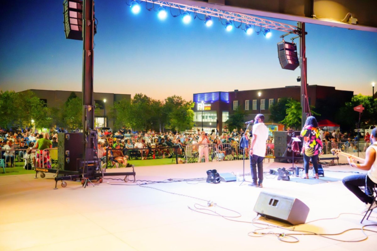 Outdoor concert at dusk with a band performing on stage and a large crowd seated beyond the barriers.