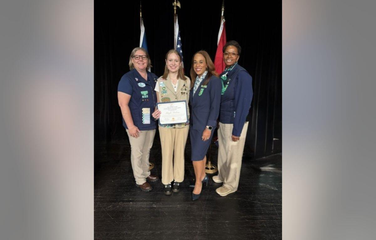 Four women stand together on a stage, one holding a certificate, with flag backdrops behind them.