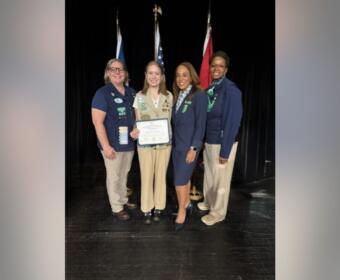 Four women stand together on a stage, one holding a certificate, with flag backdrops behind them.