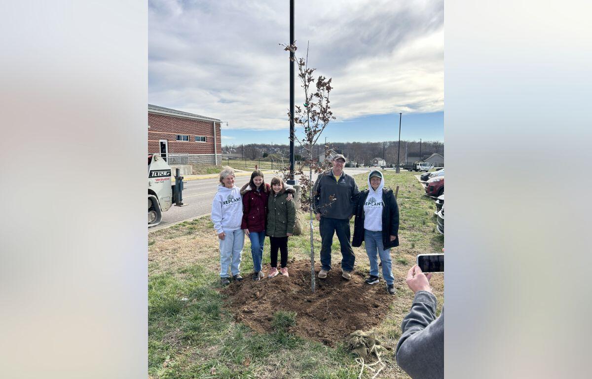 Six people stand around a newly planted sapling in a vacant lot, posing for a photo as someone records with a phone.