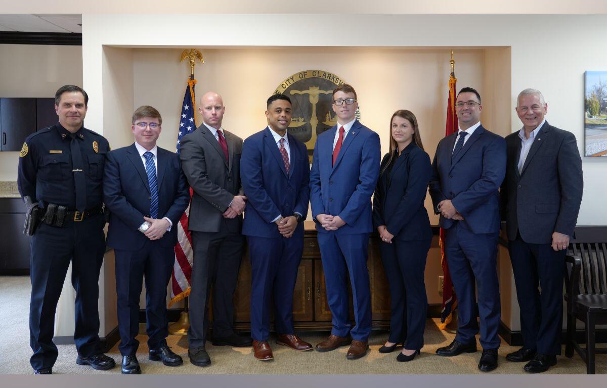 Group of eight professionals in formal attire posing in front of the City of Clarksville seal with American and state flags in a government office.