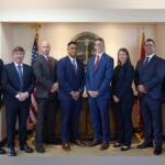 Group of eight professionals in formal attire posing in front of the City of Clarksville seal with American and state flags in a government office.