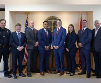 Group of eight professionals in formal attire posing in front of the City of Clarksville seal with American and state flags in a government office.
