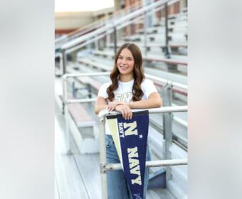 Smiling young woman leaning on a railing with a navy pennant hanging over the rail in a stadium setting.