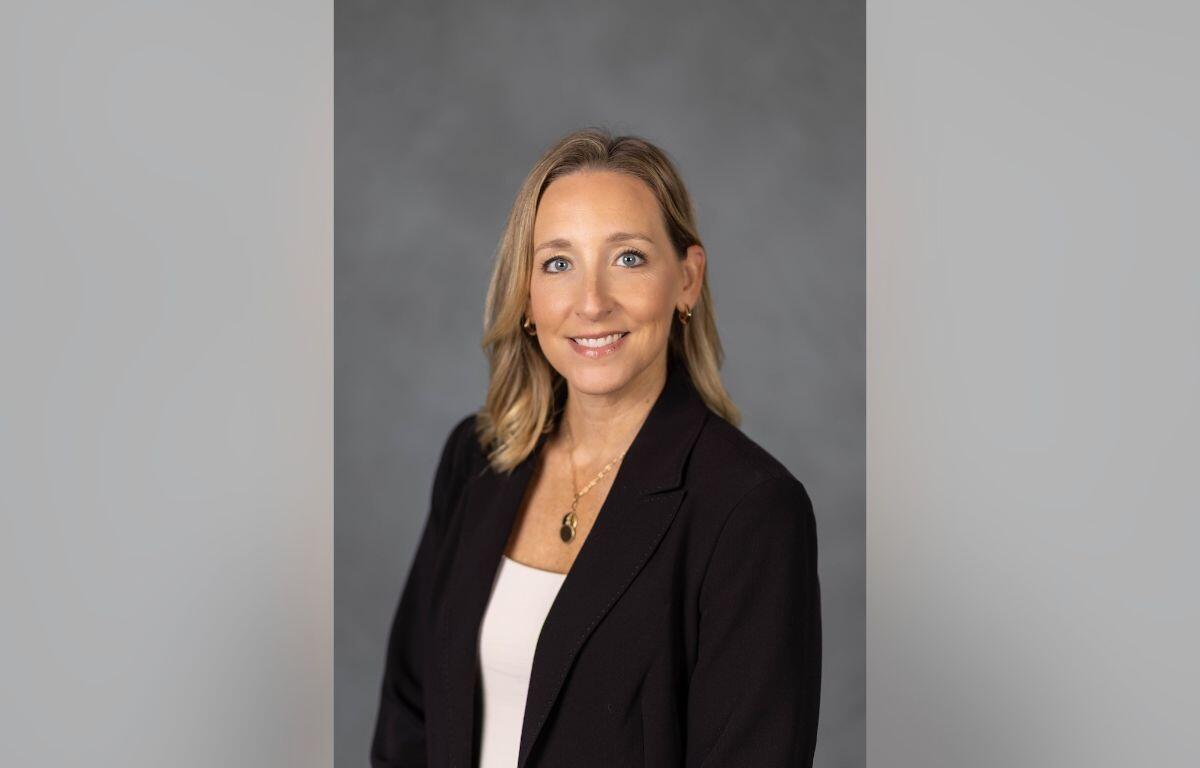 Professional headshot: smiling woman with blonde hair in a black blazer and white top, against a gray backdrop for a bio badge.