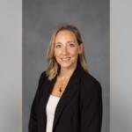 Professional headshot: smiling woman with blonde hair in a black blazer and white top, against a gray backdrop for a bio badge.