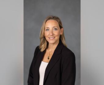 Professional headshot: smiling woman with blonde hair in a black blazer and white top, against a gray backdrop for a bio badge.